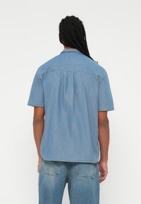 Cămiză din denim cu mânecă scurtă, de culoare albastru deschis, având guler, detaliu cu pliere la spate și o margine dreaptă. Fabrică din bumbac moale și ușor.