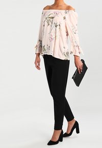 Blusa floreale off-the-shoulder color rosa pallido; pantaloni neri slim-fit; décolleté nere a punta; borsa a tracolla con un'accanto circolare.