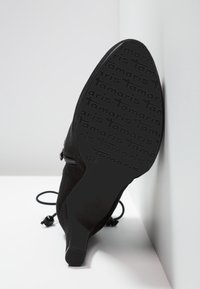 Botte noire à bout pointu avec semelle à motif portant l'inscription "Tamaris", fermeture éclair latérale et lacets décoratifs, inclinée contre un mur blanc.