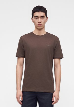 Ung mand med kort sort hår iført en ensfarvet brun Hugo t-shirt og mørke bukser, stående mod en hvid baggrund.