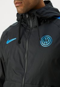 Mand iført sort zip-up jakke med blå Nike-logo og Inter Milan fodboldklubemblem på brystet.
