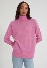 Femme aux longs cheveux bouclés portant un pull à col roulé rose et un jean bleu clair, se tenant avec les mains derrière le dos.