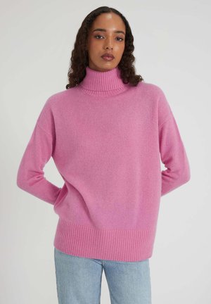 Donna con lunghi capelli ricci che indossa un maglione a collo alto rosa e jeans azzurri chiari, in piedi con le mani dietro la schiena.