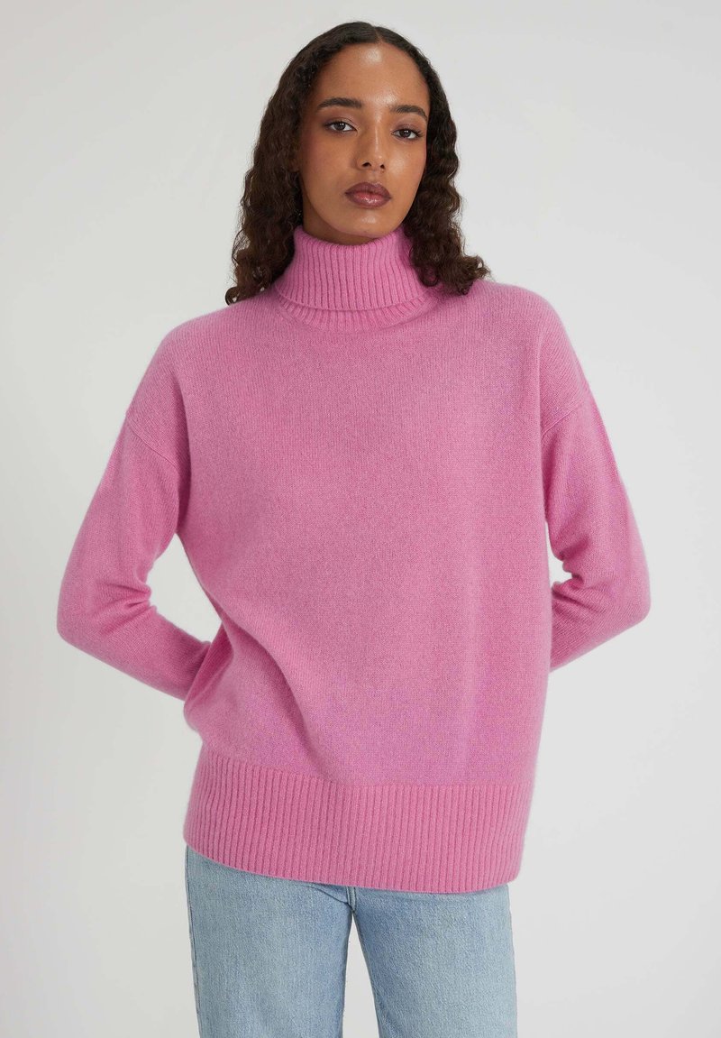 Femme aux longs cheveux bouclés portant un pull à col roulé rose et un jean bleu clair, se tenant avec les mains derrière le dos.