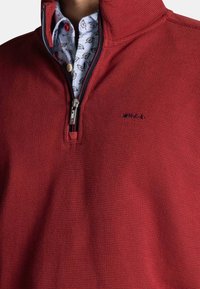 Rode ritstrui met textuurstof, voorzien van een staande kraag. Daaronder is een patroonshirt zichtbaar met lichtblauwe en bloemige ontwerpen.