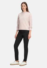 Frau steht mit einem hellrosa strukturierten Langarmoberteil, schwarzen Slim-Fit-Hosen und weißen Sneakers mit Gummisohlen vor weißem Hintergrund.