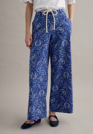 Mujer con pantalones azules de pierna ancha con estampado, cinturón de cuerda blanco, camiseta blanca con mangas de encaje y bailarinas negras con ribete azul.