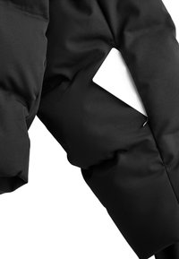 Crna podstavljena jakna s glatkim materijalom, elegantnim dizajnom i bočnom džepom u kojem se vidi bijeli predmet. Minimalistička estetika s suptilnim šavovima.