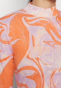 Nahaufnahme eines gerippten, langärmeligen Rollkragenoberteils mit einem abstrakten Muster aus orangefarbenen und lavendelfarbenen Wirbeln.