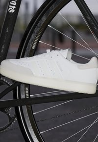 Bílé tenisky s hladkým koženým svrškem, perforovanými detaily, semišovými akcenty a texturovanou gumovou podrážkou, umístěné proti rámu bicyklu.