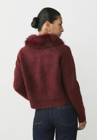 Bordeaux cropped sweater met een donzige kraag, gestructureerd breisel en geribbelde zoom. Gecombineerd met donkere spijkerbroek, weergegeven vanaf de achterkant.