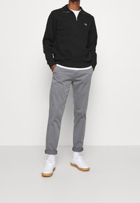 Maglione nero con zip a un quarto e logo Lacoste, abbinato a pantaloni chino grigi e sneaker bianche con suole in gomma. Design semplice e moderno.