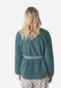 Türkisfarbener Fleece-Jacke mit weicher Textur, mit einem karierten Muster tailliert, kombiniert mit floralem Schlafanzughosen, von hinten betrachtet.