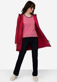 Veste imperméable rouge avec fermeture éclair intégrale, accents noirs, portée sur un haut rayé rouge et blanc. Un jean en denim noir et des baskets blanches complètent le look.