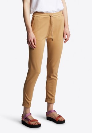 Schmal geschnittene beige Hose mit Kordelzugbund und aufgerollten Säumen, getragen mit lila und orangefarbenen Plateau-Sandalen bei einer stehenden Person.