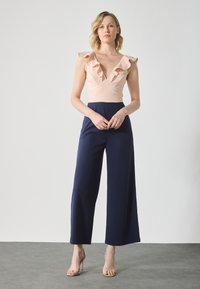 Svijetloružičasti bez rukava bodice s volanima, u kombinaciji s tamnoplavim širokim hlačama. Glatka tekstura tkanine, minimalistički dizajn i suptilni detalji.