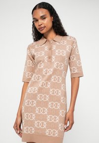 Vestido de polo de malha em bege com uma textura padronizada, apresentando logótipos de cores claras, mangas curtas e colarinho com botões.