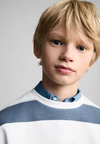 Maglione bianco con strisce blu sopra una camicia a colletto blu. Il bambino ha i capelli corti, biondo chiaro e occhi blu. Sfondo neutro.