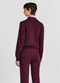 Maglione a maglia bordeaux con colletto alto, spalle scese e orlo a coste, abbinato a pantaloni aderenti coordinati. Texture liscia, design casual.