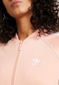 Světlounce růžová bunda na zip z hladké látky, s bílými lemovkami a malým bílým logem na levé straně hrudi.