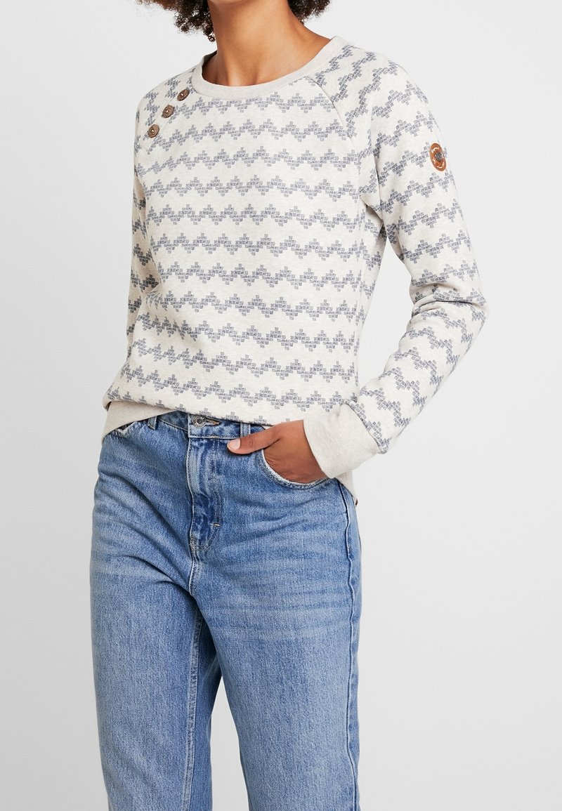 Femme portant un pull crème à manches longues avec un motif géométrique gris, des boutons en bois près de l'épaule gauche et un jean bleu clair, main dans la poche.