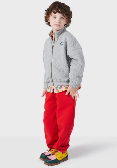 Giacca grigia con zip e colletto alto, pantaloni della tuta rossi e scarpe multicolori con accenti rosa e gialli. Camicia a fantasia visibile sotto.