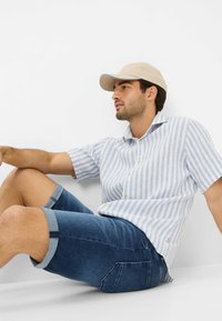 Chemise à rayures bleu clair et blanche à manches courtes avec un col, assortie à un short en denim bleu foncé. Une casquette beige avec un accent bleu sur le bord.