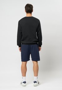 Hombre de pie de espaldas, vistiendo una camisa de manga larga negra, pantalones cortos azul marino, calcetines blancos y zapatillas blancas, sobre un fondo liso.