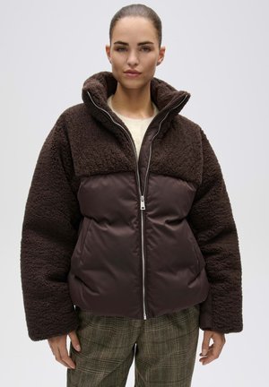 Femme portant une doudoune marron foncé zippée avec des manches et des épaules en polaire, associée à un pantalon marron à carreaux sur un fond uni.