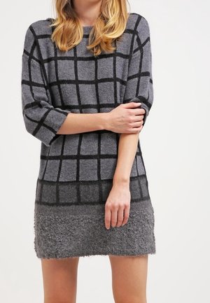 Femme portant une robe-pull grise avec des motifs en grille noire, tenant un bras croisé sur son corps devant un fond clair uni.