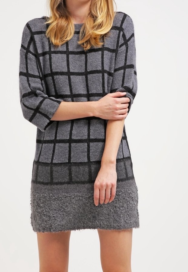 Femme portant une robe-pull grise avec des motifs en grille noire, tenant un bras croisé sur son corps devant un fond clair uni.