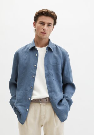 Giovane uomo con le mani in tasca, indossa una camicia azzurra con bottoni sopra una T-shirt bianca e pantaloni beige con una cintura taupe.