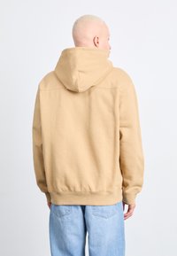 Tan hooded sweatshirt made of soft fabric, featuring ribbed cuffs and hem. Shown from behind, paired with light blue jeans.