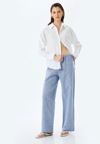 Witte button-down shirt met een losse pasvorm, lichtblauwe pantalon met wijde pijpen en een elastische tailleband, en beige teenslippers. Glad textuur van de stof.