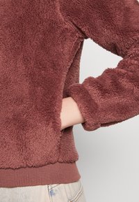 Fleecowy sweter w stonowanym kolorze różu z miękką, teksturowaną powierzchnią. Posiada ściągacz u dołu i na mankietach, luźny krój oraz kieszeń po bokach.