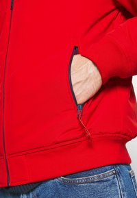 Rød zip-up jakke med en glat tekstur, der har en sidelomme med en kontrastfarvet marineblå lynlås og en snoretræk-detalje.