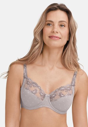 Frau mit langen blonden Haaren, die einen hellgrauen Spitzen- und Netz-BH mit bestickten Blumenmustern trägt und leicht lächelt vor weißem Hintergrund.