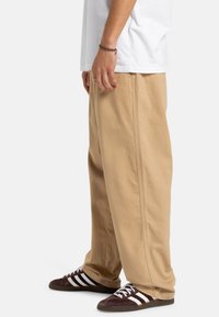 Beige wijde broek van katoen, met een zachte textuur en subtiele stikdetails, gecombineerd met donkere schoenen met witte strepen.