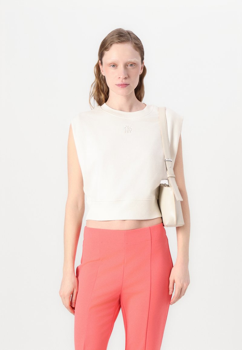 Krämfärgad kortärmad topp med präglad logotyp, matchad med högmidjade rosa byxor. Modellen håller en liten beige axelväska.