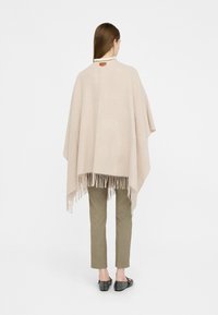 Poncho beige con orlo frangiato, realizzato in morbido tessuto. Presenta un motivo sottile e un dettaglio in pelle sul colletto. Abbinato a pantaloni beige.