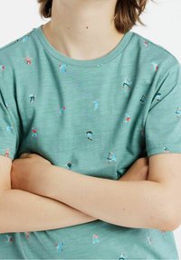 Henkilö, jolla on kädet ristissä, pukeutuneena vihreään t-paitaan, jossa on pieniä, värikkäitä kuvastoja ihmisistä pelaamassa erilaisia urheilulajeja.