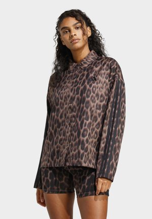 Donna con capelli ricci che indossa una giacca marrone con stampa leopardata chiusa da bottoni e pantaloncini abbinati con strisce nere sulle maniche e sui lati.