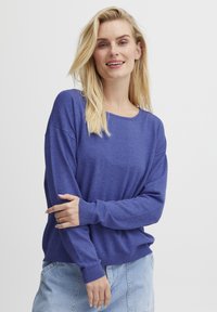 Pulover lung cu manșon purpuriu și guler rotund, cu manșete răsucite, purtat peste o fustă din denim, prezentând o croială lejeră și o textură moale.