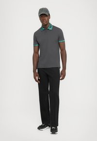 Grå polo med et mønstret turkist krage og ermeaksenter, kombinert med svarte jeans og svarte joggesko. Hvit bakgrunn.