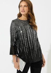 Top nero con paillettes e strisce verticali argento, scollatura tonda e maniche a campana. Abbinato a una pochette argento e pantaloni neri.