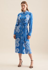 Abito camicia blu con motivo e maniche lunghe, con cintura a fiocco. Presenta una parte anteriore con bottoni, disegni floreali e lunghezza media fino al polpaccio. Sandali neri con tacco.