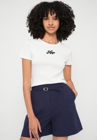 Balta kokvilnas T-kreklu ar melnu "Hugo" logotipu uz krūtīm, kombinēts ar tumši ziliem šortiem ar sprādzes detaļu jostasvietā.