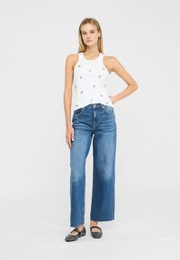 Bela majica brez rokavov z majhnimi vezenimi poudarki, kombinirana z modrimi kavbojkami širokih nogavic in visokim pasom. Črne ravne čevlje dopolnjujejo videz.