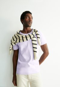 Lila ronde hals t-shirt gecombineerd met een crèmekleurige en zwarte gestreepte sweater over de schouders. Lichte broek. Glad textuur.