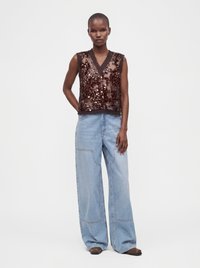 Femme portant un top marron transparent sans manches à sequins et un jean bleu clair à jambes larges, debout devant un fond blanc uni.